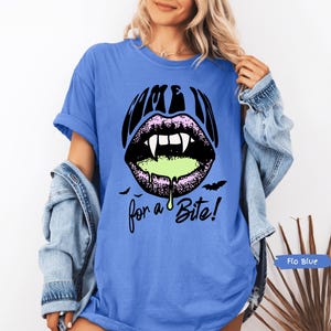 May include: A blue short-sleeve t-shirt featuring a graphic of black lips with white fangs and dripping neon green liquid. The text above the lips reads COME IN and the text below reads for a Bite! with two small black bats on either side.