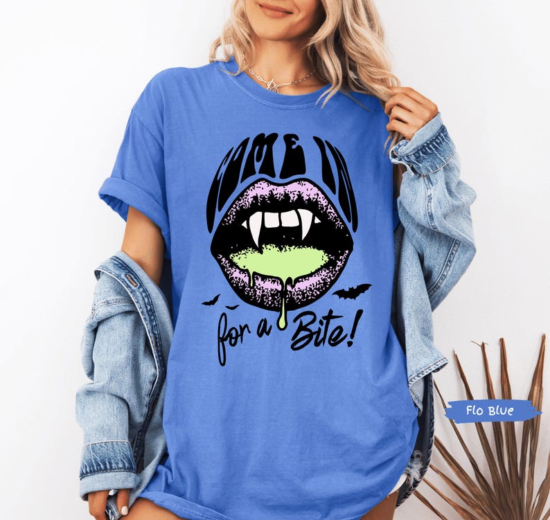 May include: A blue short-sleeve t-shirt featuring a graphic of black lips with white fangs and dripping neon green liquid. The text above the lips reads COME IN and the text below reads for a Bite! with two small black bats on either side.