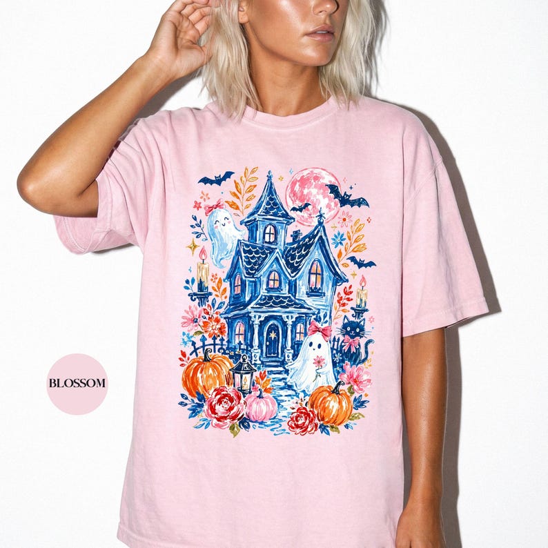 K&ouml;nnte beinhalten: Ein hellrosa T-Shirt mit einem Grafikdruck eines blauen Spukhauses, umgeben von orangefarbenen K&uuml;rbissen, roten Blumen, zwei wei&szlig;en Geistern mit rosa Schleifen, einer schwarzen Katze, Flederm&auml;usen und einem Vollmond. Das Wort BLOSSOM steht in einem Kreis.