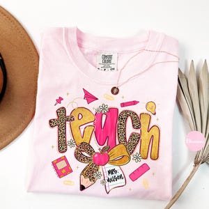 May include: A light pink Comfort Colors t-shirt with a colorful "teach" graphic. The word "teach" is in a mix of leopard print and gold lettering, with school-themed illustrations like an apple, pencil, and paper airplane.
