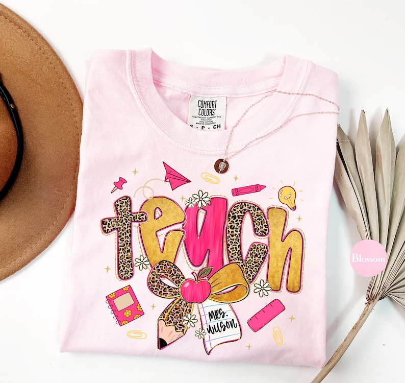 May include: A light pink Comfort Colors t-shirt with a colorful "teach" graphic. The word "teach" is in a mix of leopard print and gold lettering, with school-themed illustrations like an apple, pencil, and paper airplane.