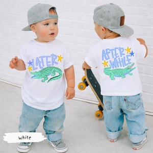 Puede incluir: Camiseta blanca para ni&ntilde;o peque&ntilde;o con un gr&aacute;fico de un cocodrilo verde y la palabra "LATER" en azul en la parte delantera. La parte trasera de la camiseta dice "AFTER WHILE" con un gr&aacute;fico de cocodrilo a juego. El ni&ntilde;o lleva vaqueros azules claros y una gorra de b&eacute;isbol gris.