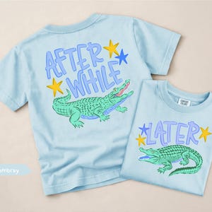 Puede incluir: Dos camisetas de color azul claro con gr&aacute;ficos de caimanes. Una camiseta tiene el texto "AFTER WHILE" y la otra dice "LATER". Ambas camisetas presentan un dise&ntilde;o de caim&aacute;n verde con estrellas amarillas y detalles azules. Las camisetas son de un material suave.