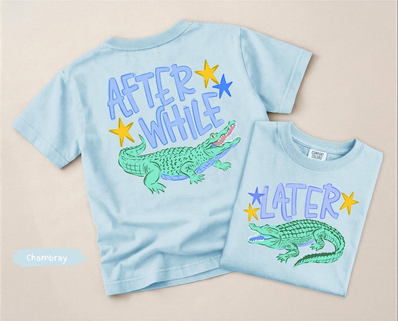 Puede incluir: Dos camisetas de color azul claro con gr&aacute;ficos de caimanes. Una camiseta tiene el texto "AFTER WHILE" y la otra dice "LATER". Ambas camisetas presentan un dise&ntilde;o de caim&aacute;n verde con estrellas amarillas y detalles azules. Las camisetas son de un material suave.