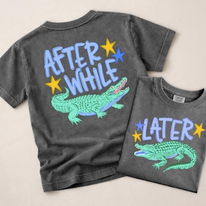 Puede incluir: Dos camisetas gris carb&oacute;n con gr&aacute;ficos de caimanes. Una camiseta dice "AFTER WHILE" con un caim&aacute;n verde y estrellas amarillas y azules. La otra camiseta dice "LATER" con un caim&aacute;n verde y estrellas amarillas y azules.