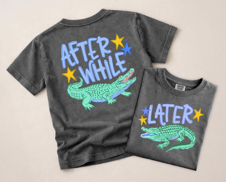 Puede incluir: Dos camisetas gris carb&oacute;n con gr&aacute;ficos de caimanes. Una camiseta dice "AFTER WHILE" con un caim&aacute;n verde y estrellas amarillas y azules. La otra camiseta dice "LATER" con un caim&aacute;n verde y estrellas amarillas y azules.