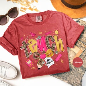 May include: Crimson-colored t-shirt with the word "TEACH" in a playful design. The letter "T" has a leopard print pattern, and the shirt features school-themed graphics like an apple, pencil, and paper airplane.
