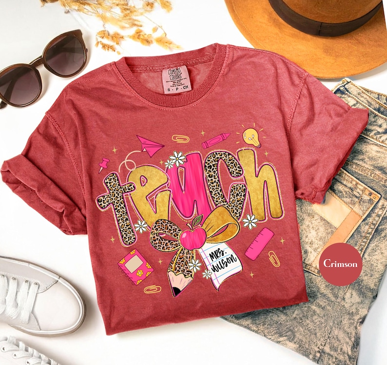 May include: Crimson-colored t-shirt with the word "TEACH" in a playful design. The letter "T" has a leopard print pattern, and the shirt features school-themed graphics like an apple, pencil, and paper airplane.