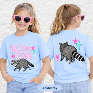 May include: Light blue t-shirts with a raccoon graphic and the text "See Ya Soon". The raccoon is gray and black with a striped tail. Pink and teal stars are also on the shirt.
