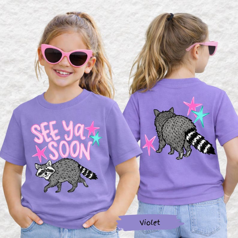 May include: A violet t-shirt featuring a raccoon graphic and the text "See Ya Soon" in pink and teal. The back of the shirt also has a raccoon graphic and star accents. The shirt is made of a soft material.