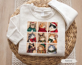 May include: A natural-colored crewneck sweatshirt featuring a graphic print of nine individual cat illustrations. Each cat is depicted in a festive holiday theme, wearing items such as Santa hats, scarves, reindeer antlers, and Christmas lights.