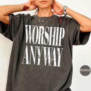 Puede incluir: Una camiseta de manga corta gris carbón con el texto blanco desgastado "WORSHIP ANYWAY" impreso en la parte delantera. La camiseta tiene cuello redondo y un corte holgado. Un pequeño icono circular en la esquina inferior derecha muestra la palabra "PEPPER".