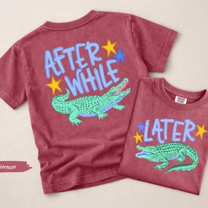 Puede incluir: Dos camisetas carmes&iacute; con gr&aacute;ficos de caimanes. Una camiseta dice "AFTER WHILE" con un caim&aacute;n verde y estrellas amarillas y azules. La otra camiseta dice "LATER" con un caim&aacute;n verde y estrellas amarillas y azules.