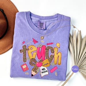 May include: A violet Comfort Colors t-shirt with a graphic that says "Teach" in a mix of fonts and colors. The design includes a pencil, apple, and other school-themed elements. A gold necklace is draped across the shirt.