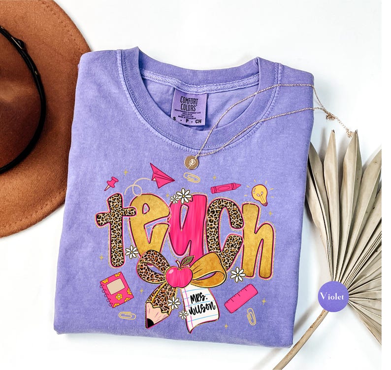 May include: A violet Comfort Colors t-shirt with a graphic that says "Teach" in a mix of fonts and colors. The design includes a pencil, apple, and other school-themed elements. A gold necklace is draped across the shirt.