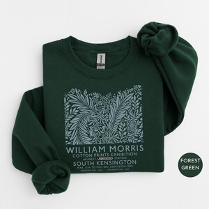 Puede incluir: Sudadera verde bosque con un dise&ntilde;o floral y texto de la exposici&oacute;n William Morris Cotton Prints. El texto incluye la ubicaci&oacute;n, South Kensington, y las fechas del 12 de marzo al 18 de mayo. La sudadera tiene mangas largas.