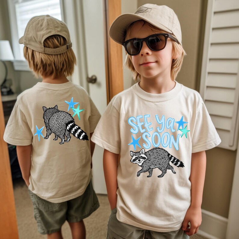 May include: Beige t-shirts with a raccoon graphic and the text "See Ya Soon". The raccoon is gray and white with a striped tail. The text and stars are light blue.