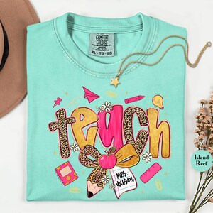 May include: A turquoise Comfort Colors t-shirt with a colorful "Teach" graphic. The word "Teach" is designed with leopard print and pink accents, along with school-themed illustrations like an apple, pencil, and notebook. A gold necklace with a star pendant is draped on the shirt.