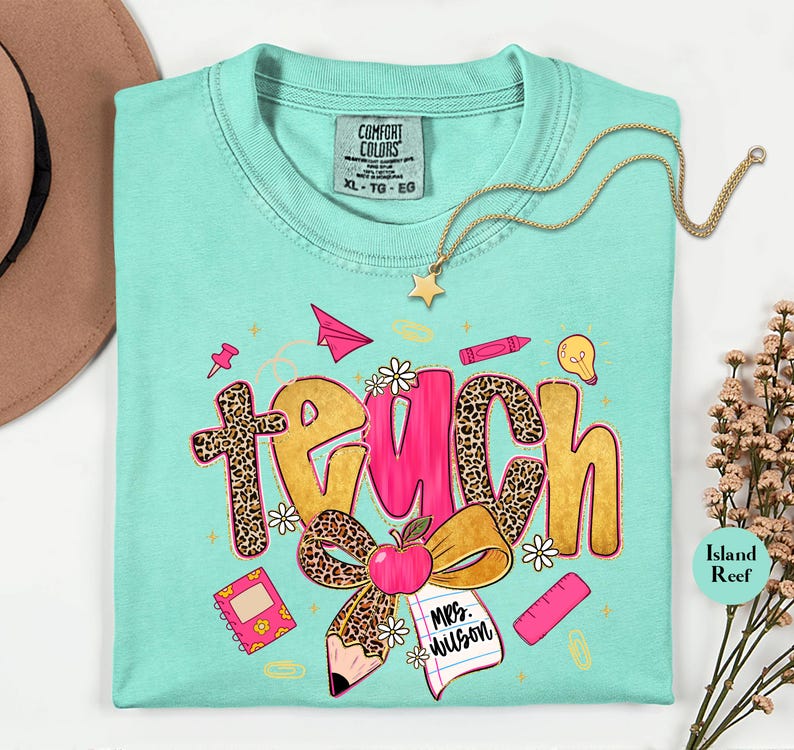May include: A turquoise Comfort Colors t-shirt with a colorful "Teach" graphic. The word "Teach" is designed with leopard print and pink accents, along with school-themed illustrations like an apple, pencil, and notebook. A gold necklace with a star pendant is draped on the shirt.