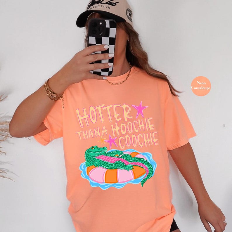 Puede incluir: Camiseta color mel&oacute;n ne&oacute;n con el texto "Hotter Than A Hoochie Coochie" y un gr&aacute;fico de un caim&aacute;n verde sobre una boya rosa y naranja. La camiseta tiene mangas cortas y un corte holgado.