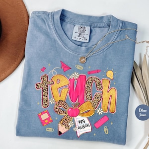 May include: Blue jean Comfort Colors t-shirt with a graphic that says "Teach". The word "Teach" is in a combination of leopard print, pink, and gold. The graphic includes an apple, pencil, and other school-themed elements.