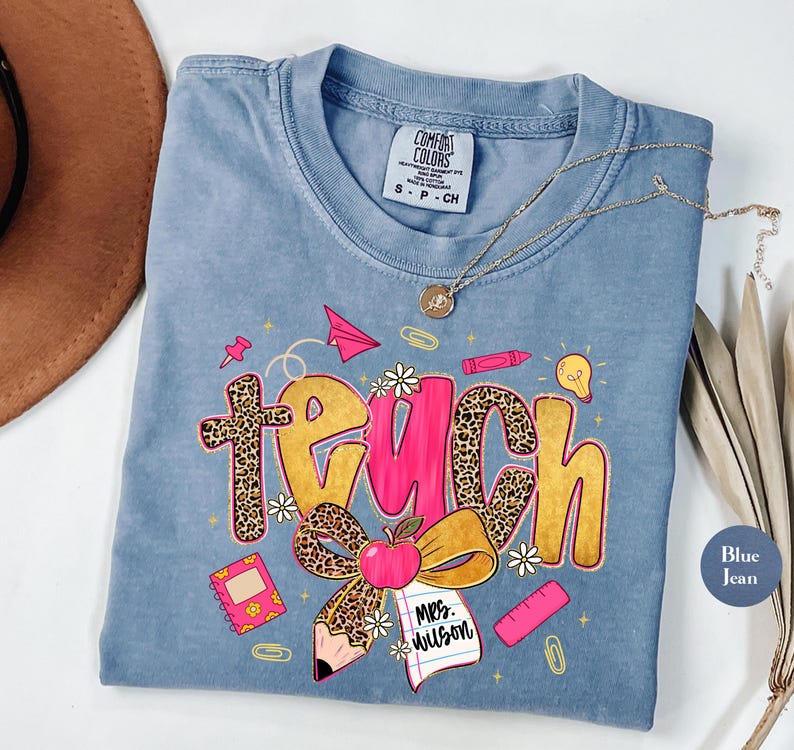 May include: Blue jean Comfort Colors t-shirt with a graphic that says "Teach". The word "Teach" is in a combination of leopard print, pink, and gold. The graphic includes an apple, pencil, and other school-themed elements.