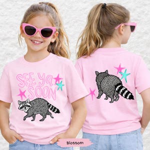 May include: Pink short-sleeved t-shirt with a raccoon graphic and the text "See Ya Soon". The raccoon is gray with a black and white striped tail. The design includes pink and teal stars.