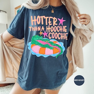 Puede incluir: Camiseta azul marino con la frase "Hotter Than a Hoochie Coochie" en rosa y naranja. Un cocodrilo de dibujos animados flota en una boya. La camiseta es de corte holgado.
