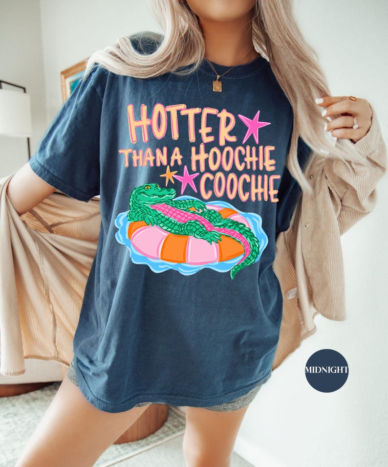 Puede incluir: Camiseta azul marino con la frase "Hotter Than a Hoochie Coochie" en rosa y naranja. Un cocodrilo de dibujos animados flota en una boya. La camiseta es de corte holgado.