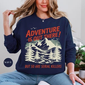 K&ouml;nnte beinhalten: Marineblaues Sweatshirt mit Berg- und Waldmotiv. Der Text lautet "ADVENTURE IS OUT THERE! BUT SO ARE SERIAL KILLERS" in Rot und Wei&szlig;. Das Sweatshirt hat einen Rundhalsausschnitt und lange &Auml;rmel.