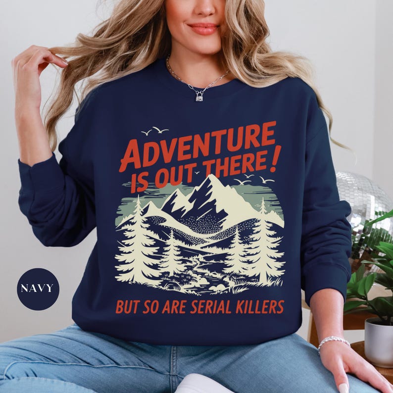 K&ouml;nnte beinhalten: Marineblaues Sweatshirt mit Berg- und Waldmotiv. Der Text lautet "ADVENTURE IS OUT THERE! BUT SO ARE SERIAL KILLERS" in Rot und Wei&szlig;. Das Sweatshirt hat einen Rundhalsausschnitt und lange &Auml;rmel.