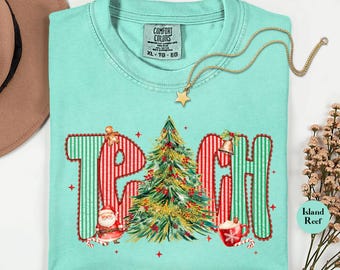 May include: A light teal Comfort Colors XL t-shirt with the word "TEACH" in a Christmas-themed design. The letters are red and green with festive illustrations of a Christmas tree, Santa, and a mug of hot cocoa. A gold star necklace is draped across the shirt.