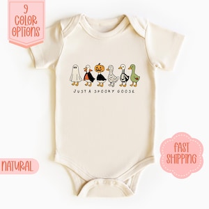 May include: A cream-coloured baby bodysuit featuring a Halloween-themed graphic. The design shows cartoon geese dressed as a ghost, a witch, a pumpkin, a mummy, and a zombie. Text reads "JUST A SPOOKY GOOSE".