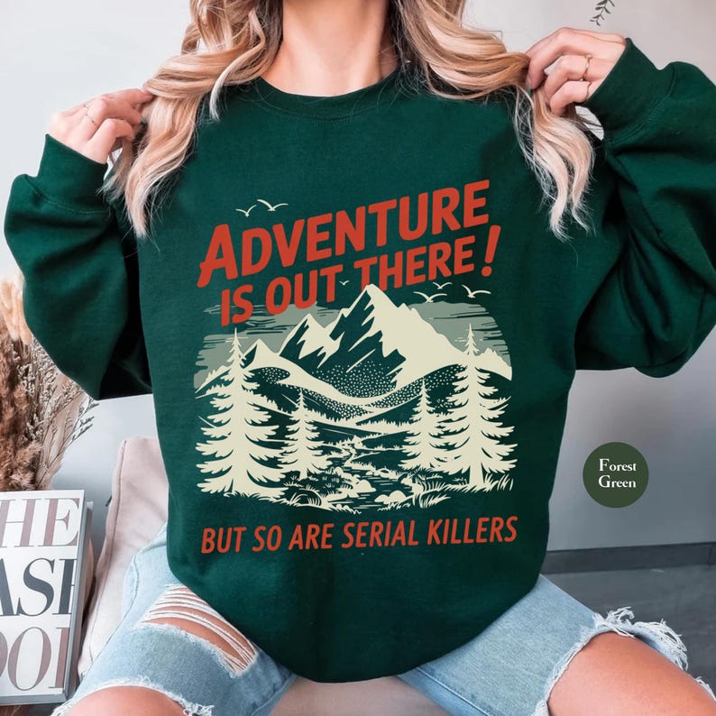 K&ouml;nnte beinhalten: Dunkelgr&uuml;nes Sweatshirt mit dem Text "ADVENTURE IS OUT THERE! BUT SO ARE SERIAL KILLERS" in Rot und Wei&szlig;. Die Grafik zeigt eine Bergkette, B&auml;ume und V&ouml;gel. Das Sweatshirt hat einen Rundhalsausschnitt.