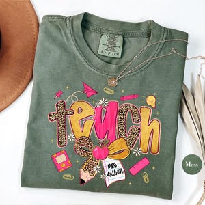May include: A moss green t-shirt with the word "teach" in a playful design. The word "teach" features a leopard print cross and bow, with a pink center. The shirt includes school-themed graphics like a pencil, apple, and notebook.