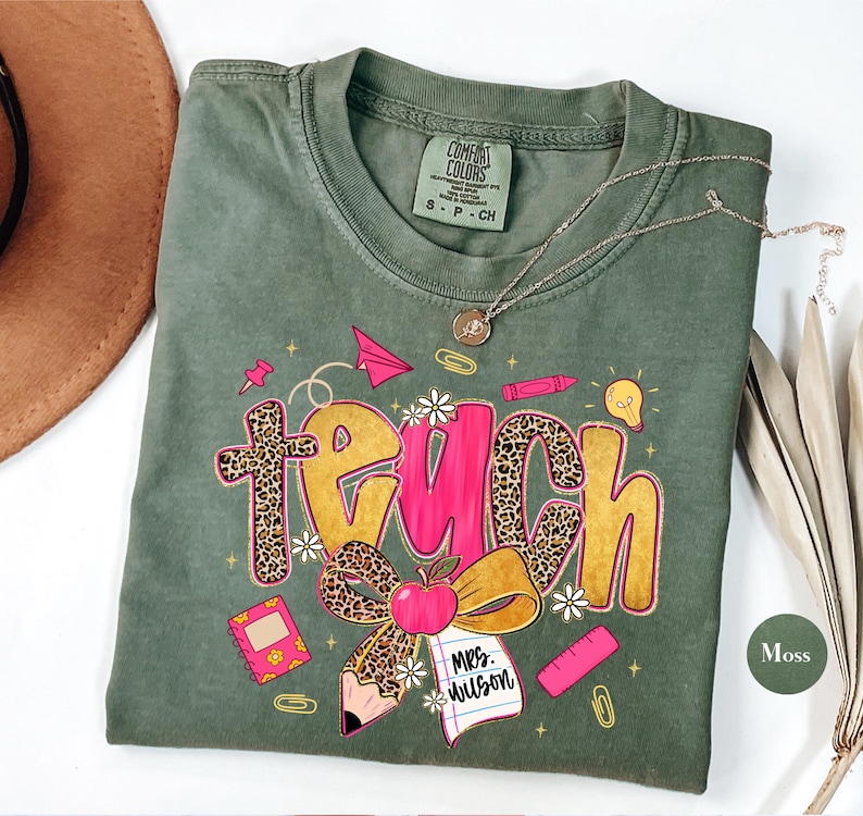 May include: A moss green t-shirt with the word "teach" in a playful design. The word "teach" features a leopard print cross and bow, with a pink center. The shirt includes school-themed graphics like a pencil, apple, and notebook.