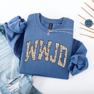 May include: Indigo blue sweatshirt with the letters WWJD in a camouflage pattern. A silver necklace with a small pendant rests on the sweatshirt. The sweatshirt is paired with light blue ripped jeans.