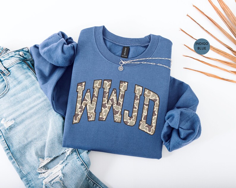 May include: Indigo blue sweatshirt with the letters WWJD in a camouflage pattern. A silver necklace with a small pendant rests on the sweatshirt. The sweatshirt is paired with light blue ripped jeans.
