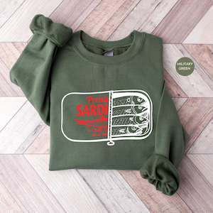 May include: Military green sweatshirt featuring a sardine can graphic. The graphic includes the words "Premium Sardines" in red, with a white outline of a sardine can containing three fish.