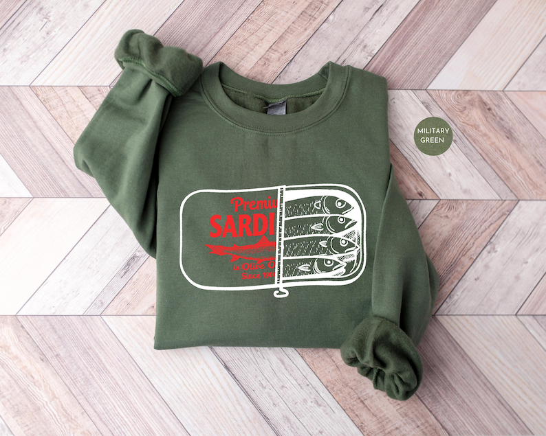 May include: Military green sweatshirt featuring a sardine can graphic. The graphic includes the words "Premium Sardines" in red, with a white outline of a sardine can containing three fish.