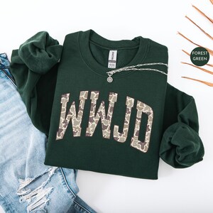 May include: A forest green sweatshirt with the letters "WWJD" in a camouflage pattern. A silver necklace with a pendant rests on the sweatshirt. The sweatshirt is paired with blue jeans.