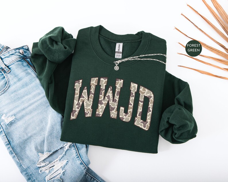 May include: A forest green sweatshirt with the letters "WWJD" in a camouflage pattern. A silver necklace with a pendant rests on the sweatshirt. The sweatshirt is paired with blue jeans.