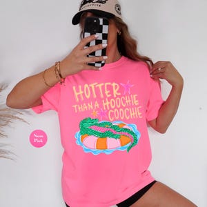 Puede incluir: Camiseta rosa ne&oacute;n con el texto amarillo "HOTTER THANA HOOCHIE COOCHIE" y un caim&aacute;n verde sobre una boya naranja y azul. La camiseta es de manga corta.
