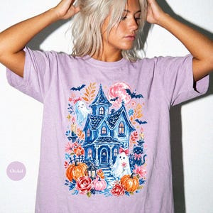 K&ouml;nnte beinhalten: Ein hellviolettes Kurzarm-T-Shirt mit einem Grafikdruck eines blauen Spukhauses, zwei Geistern, einer schwarzen Katze, K&uuml;rbissen, Blumen, Flederm&auml;usen und einem rosa Mond. Das Design enth&auml;lt Kerzen und florale Elemente in einer bunten Illustration.
