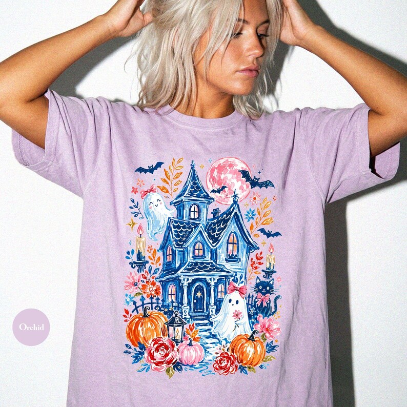 K&ouml;nnte beinhalten: Ein hellviolettes Kurzarm-T-Shirt mit einem Grafikdruck eines blauen Spukhauses, zwei Geistern, einer schwarzen Katze, K&uuml;rbissen, Blumen, Flederm&auml;usen und einem rosa Mond. Das Design enth&auml;lt Kerzen und florale Elemente in einer bunten Illustration.
