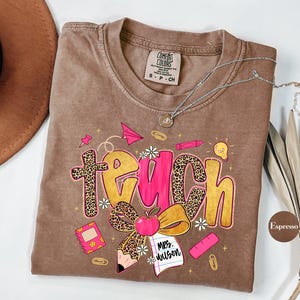 May include: Espresso-colored t-shirt with the word "TEACH" in a playful design. The letters feature leopard print and pink accents, with school-themed graphics like a pencil, apple, and paper airplane. A silver necklace rests on the shirt.