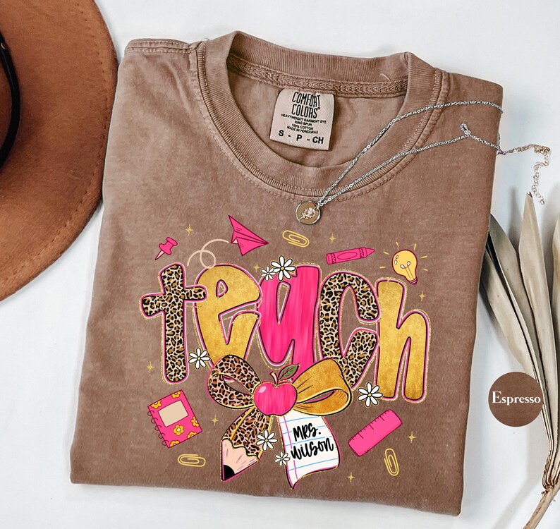 May include: Espresso-colored t-shirt with the word "TEACH" in a playful design. The letters feature leopard print and pink accents, with school-themed graphics like a pencil, apple, and paper airplane. A silver necklace rests on the shirt.