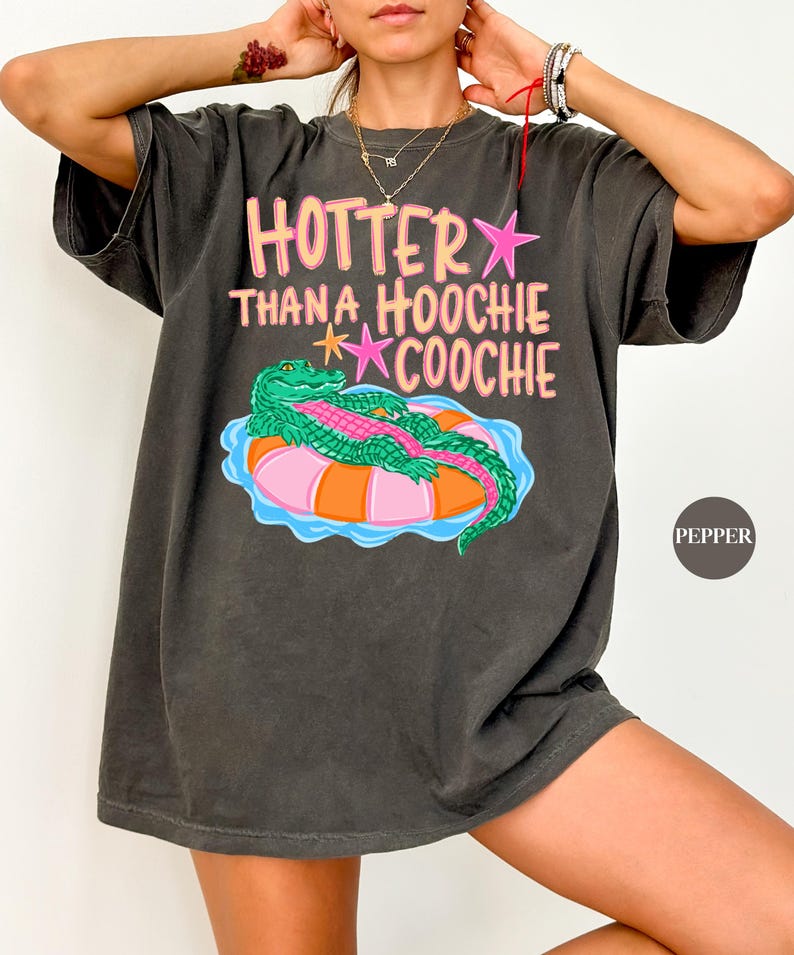 Puede incluir: Camiseta gris oscuro con el texto "Hotter Than a Hoochie Coochie" en rosa y amarillo. La camiseta presenta un gr&aacute;fico de un caim&aacute;n verde sobre un flotador inflable rosa y naranja. La camiseta es de corte holgado.