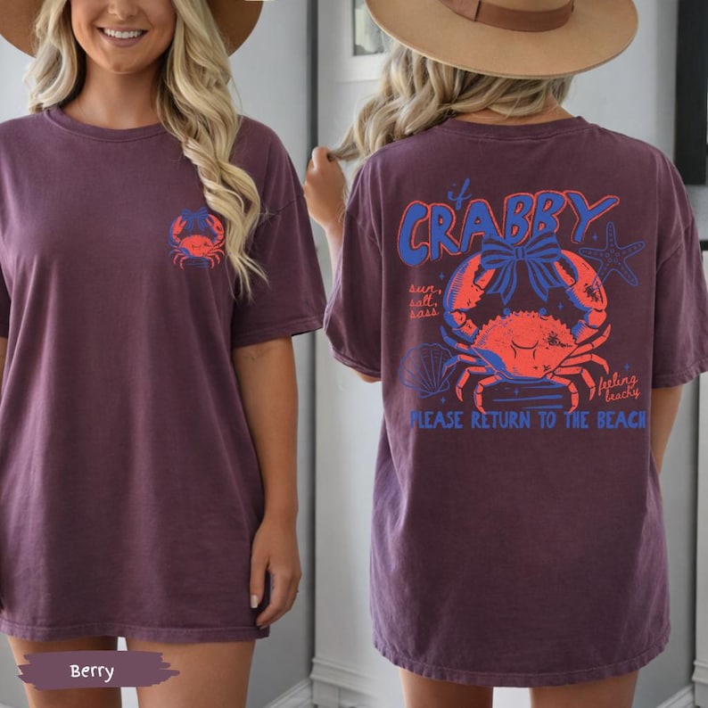 Puede incluir: Camiseta de color bayas con un gr&aacute;fico de cangrejo en la parte delantera y trasera. La parte trasera de la camiseta presenta el texto "It's Crabby" encima de una ilustraci&oacute;n de cangrejo, con las palabras "sun, salt, sass" y "feeling beachy" y "PLEASE RETURN TO THE BEACH".
