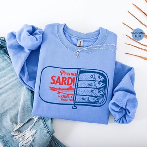 May include: Carolina blue sweatshirt with a sardine can graphic. The graphic features the text "Premium SARDI" in red, a red fish, and the text "in Olive Oil Since 1900" in black. A silver necklace is draped across the sweatshirt.